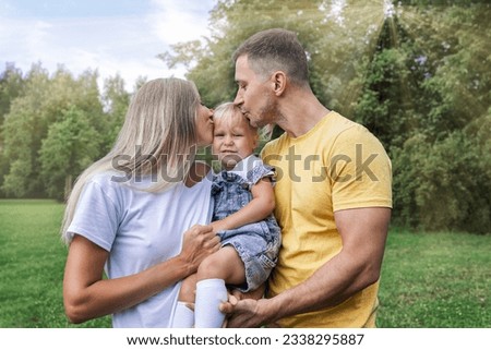 Happy parents with their little daughter in the park on a sunny summer day. Laughing young woman with a man and a child in jeans and t-shirts hug and kiss. Love and tenderness.