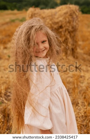 a beautiful blonde girl with long hair in a linen dress is standing by a haystack