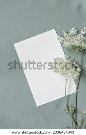 Empty paper card mock up with white flowers on a light pale blue textile background. Aesthetic minimalist wedding invitation, business brand template