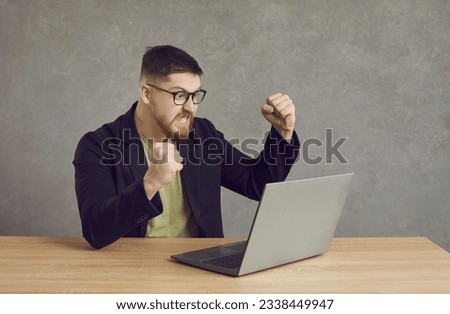 Funny man having problem with computer. Angry furious crazy young guy in glasses sitting at desk, clenching fists and looking at laptop screen threatening to hit and break it. Software error concept Royalty-Free Stock Photo #2338449947