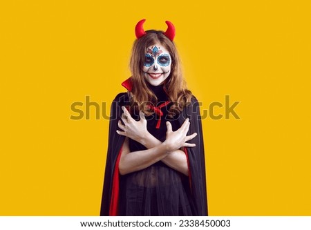 Portrait of teen girl with creative Halloween skull makeup. Beautiful teenager wearing red horns and cape posing with her arms crossed over isolated studio background with copy space
