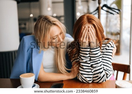 Portrait of displeased young woman and best female friend trying to comfort and cheer up sitting together in cafe by window. Unhappy redhead lady covering face with hands and crying. Royalty-Free Stock Photo #2338460859