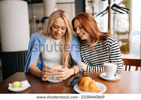 Portrait of two cheerful young women friends sitting at coffee shop having coffee and chatting sitting at table after long separation. Happy redhead and blonde female meeting at cafe on weekend. Royalty-Free Stock Photo #2338460869