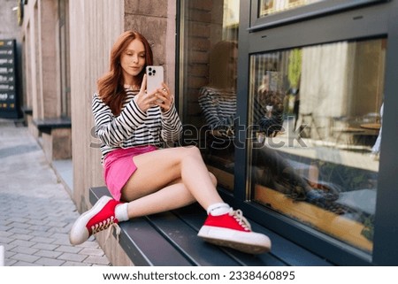 Attractive young woman looking at smartphone screen, enjoying video call or taking selfie picture outdoors, sitting by window on windowsill. Redhead female speaking in videocall on phone