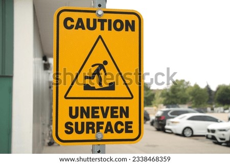 caution uneven surface sign with caption and illustration picture of person walking on an uneven surface surrounded by a triangle, building wall and parking lot with parked cars and sky in background Royalty-Free Stock Photo #2338468359
