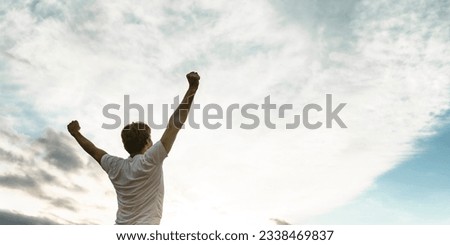 Young man standing with his arms raised high in triumph under white cloudy sky with plenty of copy space. Royalty-Free Stock Photo #2338469837