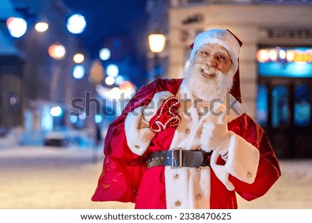Santa Claus on Xmas Eve at city streets. Friendly cheerful outgoing senior man with real white beard Father Christmas at snowy old town lifestyle scenery. New Year, celebration, garlands illumination