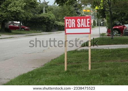 generic for sale sign on front lawn of residential home in white writing on red background and wood posts with blurred long street behind - medium shot frame right.