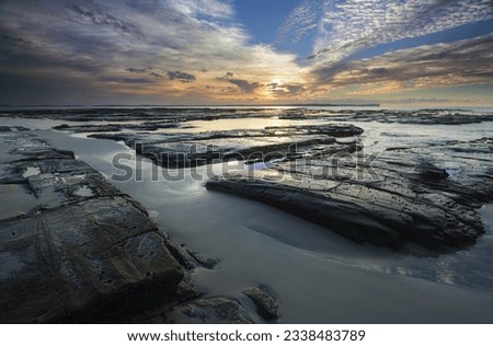 The light from the sun filtered through high cloud just after sunrise falls across the rocks wet sand and shallow tidal flows at Plantation Point, Vincentia in Jervis Bay creating interesting ares of