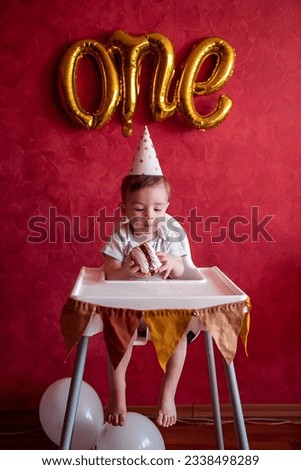Little boy of one year old in party hat sits in childrens chair, eating birthday cake, Meringue, blows out candle. Kid on red isolated background, on textured wall foil gold balloons One. Home party
