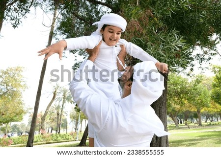 Arabian family together as father tosses his son in the air having fun at an outdoor location with trees around. Arab dad and child bonding wearing Kandura Thobe Thawb. Royalty-Free Stock Photo #2338555555