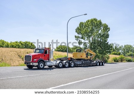 Industrial standard professional red day cab big rig heavy duty semi truck tractor transporting fastened construction equipment on step down semi trailer driving on the wide highway road Royalty-Free Stock Photo #2338556255