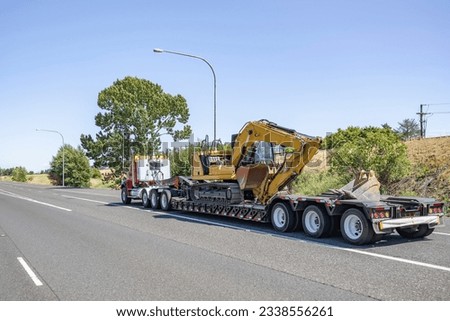Industrial standard professional red day cab big rig heavy duty semi truck tractor transporting fastened construction equipment on step down semi trailer driving on the wide highway road Royalty-Free Stock Photo #2338556261