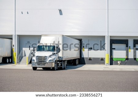 Industrial standard professional white day cab big rig heavy duty semi truck tractor with dry van semi trailer standing on the warehouse dock gate loading cargo for the next commercial delivery Royalty-Free Stock Photo #2338556271