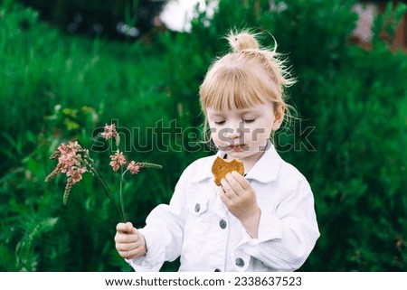 a very cute blonde girl in a white jacket is holding a pink flower and eating cookies in the park. happy childhood