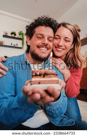 Vertical portrait of happy young couple celebrating a birthday with a little cake blowing the candle. Wife giving a funny surprise to her husband for their anniversary while hugging him from behind