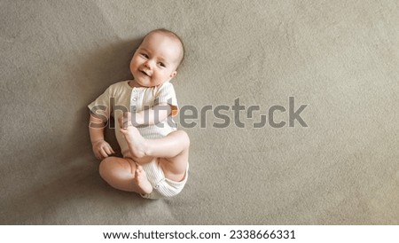 Infant lying on brown bed smiling with joy. Baby girl with delighted expression plays touching toes with hands and relaxing on bed, copyspace Royalty-Free Stock Photo #2338666331