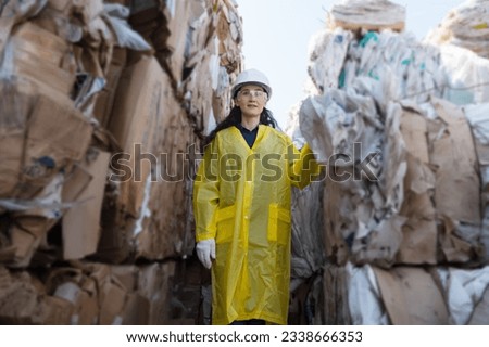 Woman supervisor checks operations in workshop of large waste recycling plant. Female in white work hat among pile of unsorted waste in plant Royalty-Free Stock Photo #2338666353