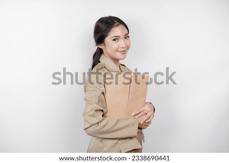 A smiling young beautiful Asian civil worker with brown uniform bringing document. Indonesian PNS. Royalty-Free Stock Photo #2338690441