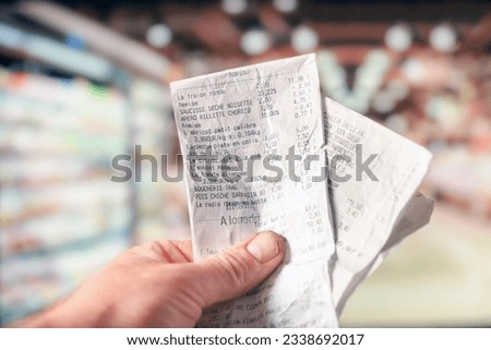 hand holding receipts in the supermarket with french list means "Hazelnut dry sausage,
Apero rillette chorizo,
Small size apricot,
Flat peach in package,
Saithe back,
Lime by the piece,
hash avocado"
 Royalty-Free Stock Photo #2338692017