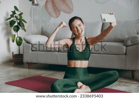 Full body young strong sporty athletic fitness trainer instructor woman in green tracksuit do selfie shot on mobile cell phone show muscles train do exercises at home gym indoor Workout sport concept