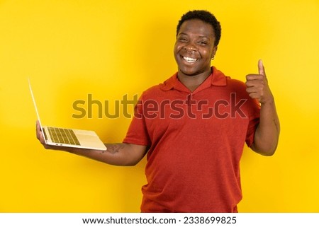 Beautiful attractive Young latin man wearing red T-shirt over yellow background hold netbook hand finger show thumb up