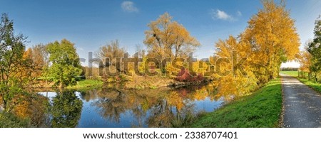 Panoramic photo of colorful autumn leaves on the bank of the river Nidda with its calm water surface in a nature park near Frankfurt am Main with a riverside path on the edge
