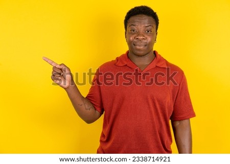 Young latin man wearing red T-shirt over yellow background points to side on blank space demonstrates advertisement. People and promotion concept