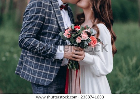 bride and groom kissing, young and beautiful couple, caucasian man and woman, dressed in suit and white wedding dress, holding bouquet with pink roses, green trees in background, cropped image