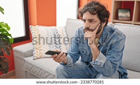 Young hispanic man watching tv sitting on sofa with boring expression at home