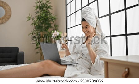 Young beautiful hispanic woman wearing bathrobe drinking champagne having video call at bedroom