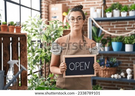 Hispanic woman working at florist holding open sign relaxed with serious expression on face. simple and natural looking at the camera. 