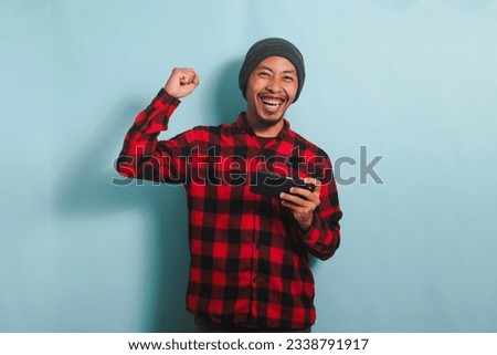 Excited young Asian man gamer with beanie hat and red plaid flannel shirt is raising his fist in a YES gesture while winning and playing an online game on his smartphone, isolated on a blue background
