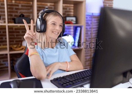 Young caucasian woman playing video games wearing headphones smiling with happy face winking at the camera doing victory sign with fingers. number two. 