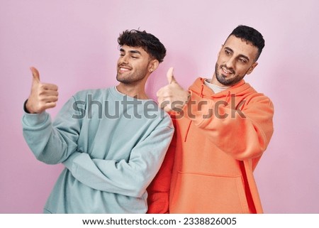 Young hispanic gay couple standing over pink background looking proud, smiling doing thumbs up gesture to the side 