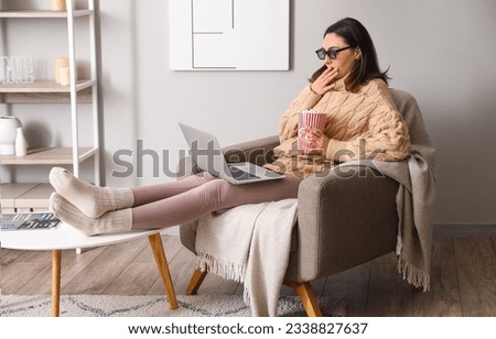 Shocked young woman with 3D glasses and popcorn watching movie in armchair at home