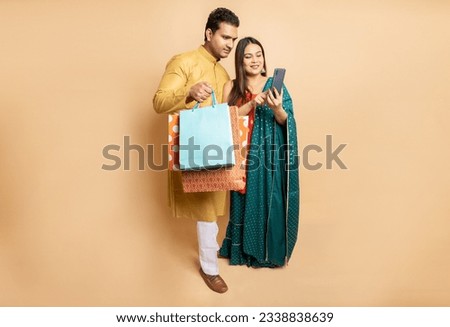 Happy young indian couple wearing traditional cloths holding shopping bags and and smart phone isolated on beige studio background. Online shopping, Diwali celebration and festive sale, copy space. Royalty-Free Stock Photo #2338838639