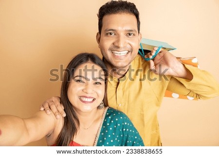 Happy young indian couple wearing traditional cloths holding shopping bags taking selife picture of their own isolated on beige background. Diwali celebration and festive offer and sale.