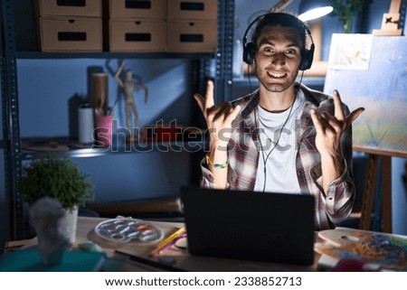 Young hispanic man sitting at art studio with laptop late at night shouting with crazy expression doing rock symbol with hands up. music star. heavy concept. 
