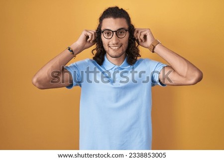 Young hispanic man standing over yellow background smiling pulling ears with fingers, funny gesture. audition problem 