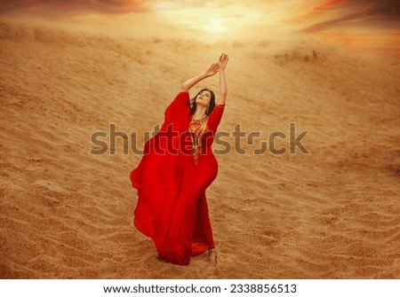 arabic woman in red long dress stands in desert long train silk fabric fly flutter in wind motion. clothes gold beauty face hand raised to sky. fashion model black hair. Sand dunes sunset sun
