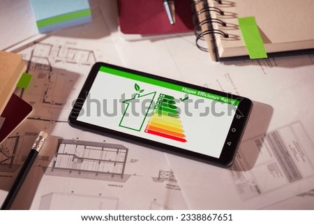 Energy efficiency screen on cellphone leaning on drawings and blueprints of a new house. Architect's desk with plans on it and a cell phone with an energy efficiency graph. Renewable energy concept. Royalty-Free Stock Photo #2338867651