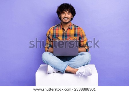 Full size photo of smart man wear checkered shirt jeans shoes sit on cube with laptop typing email isolated on purple color background