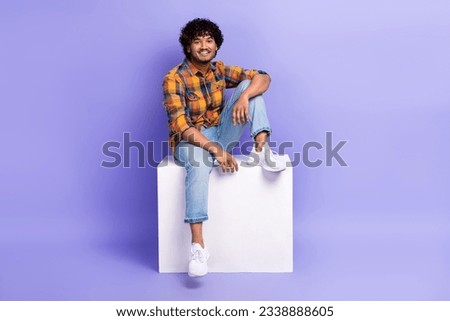 Full length photo of handsome nice man dressed plaid shirt denim pants sit on white cube platform isolated on purple color background