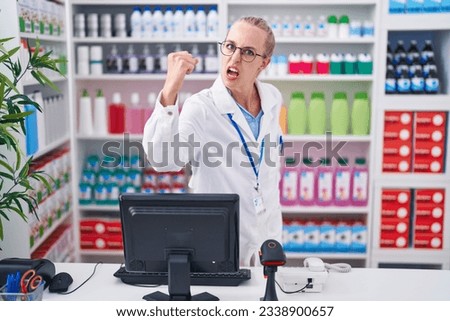 Young caucasian woman working at pharmacy drugstore angry and mad raising fist frustrated and furious while shouting with anger. rage and aggressive concept.  Royalty-Free Stock Photo #2338900657