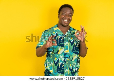 Young latin man wearing hawaiian shirt over yellow background showing both hands with fingers in OK sign. Approval or recommending concept