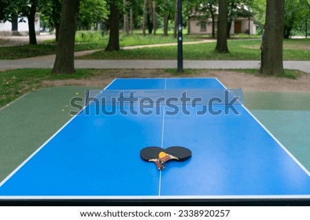 Two tennis rackets and an orange tennis ball lie on a blue tennis table next to a net in a city park ping pong game