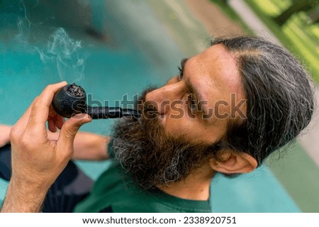 Portrait of an aged man with long hair and gray beard smoking a pipe in city park against the background of trees