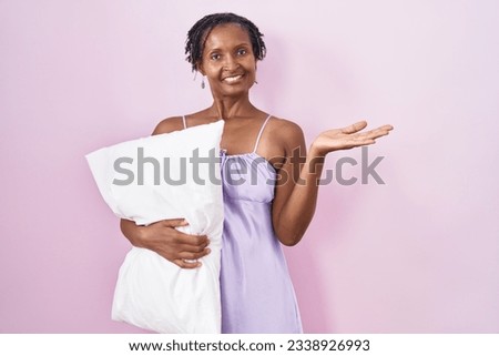 African woman with dreadlocks wearing pajama hugging pillow smiling cheerful presenting and pointing with palm of hand looking at the camera. 