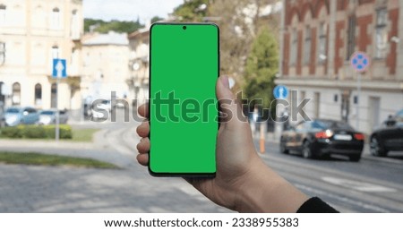 Woman Hand is holding Cell Phone outdoor on the Street in old European City. Green Screen in Vertical Position, Lot of People, Cars on the Background, good Weather outside. Apps. Smartphone.